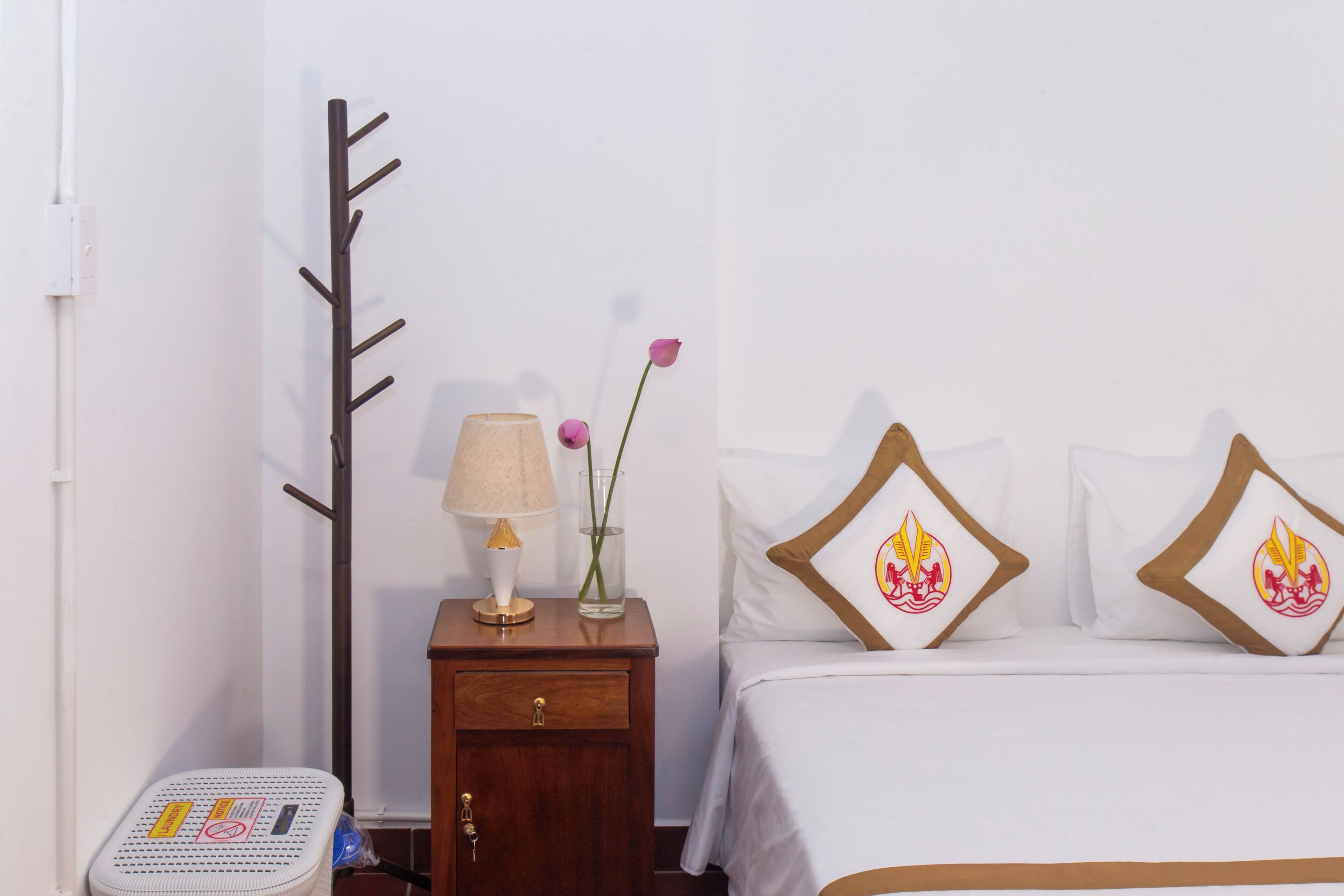 A hotel room with a bed, a nightstand with a lamp, a vase with a rose, and a coat rack.