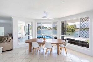 A modern dining area with a large table and chairs, large windows with a view of a pool and water body, and a ceiling fan. - Little Casa - Oceanstays - Pool & Boat Ramp (Yamba)