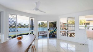 A spacious room with a large dining table, a ceiling fan, and a view of a water body and playground through large windows. - Little Casa - Oceanstays - Pool & Boat Ramp (Yamba)