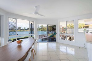A spacious room with a dining table, a ceiling fan, and large windows overlooking a water body. - Little Casa - Oceanstays - Pool & Boat Ramp (Yamba)