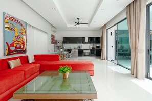 A modern living room with a red sofa, a glass coffee table, and a kitchen in the background. - 3 Bedrooms Private Pool Villa (Tambon Rawai)