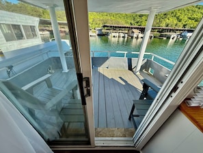 A boat deck with a glass door leading to a covered area, a wooden deck, and a view of docked boats and greenery.