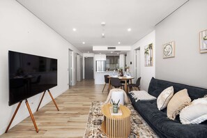 Ein modernes Wohnzimmer mit einem Flachbildfernseher, einem dunkelgrauen Sofa, einem runden Holztisch und einem gemusterten Teppich.