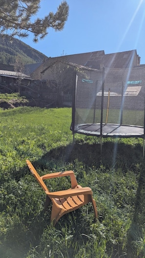 En studsmatta med skyddsnät, en trästol och en gräsyta med en byggnad i bakgrunden.