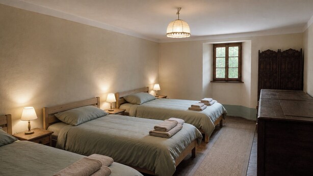A room with three beds, each with a bedside lamp and folded towels. There is a window with a view of greenery outside.