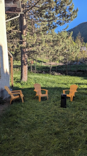 A grassy outdoor area with three wooden Adirondack chairs and a black trash bin.