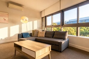 A modern living room with a sectional sofa, a wooden coffee table, and large windows with blinds. - Central 2BR Olympic Park Apt with Parking (Sydney Olympic Park)