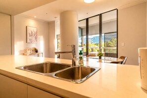 A modern kitchen with two stainless steel sinks, a large window with blinds, and a dining area with a table and chairs. - Central 2BR Olympic Park Apt with Parking (Sydney Olympic Park)