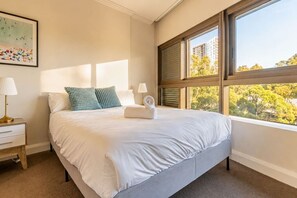 A neatly made bed with white bedding, a bedside lamp, and a window with blinds. - Central 2BR Olympic Park Apt with Parking (Sydney Olympic Park)