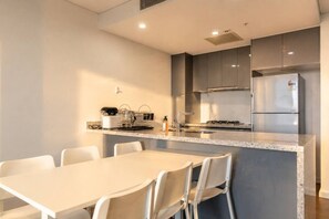 A modern kitchen with a central island, white chairs, and a stainless steel refrigerator. - Stylish 2BR Olympic Park | Pool, Gym & Parking (Sydney Olympic Park)