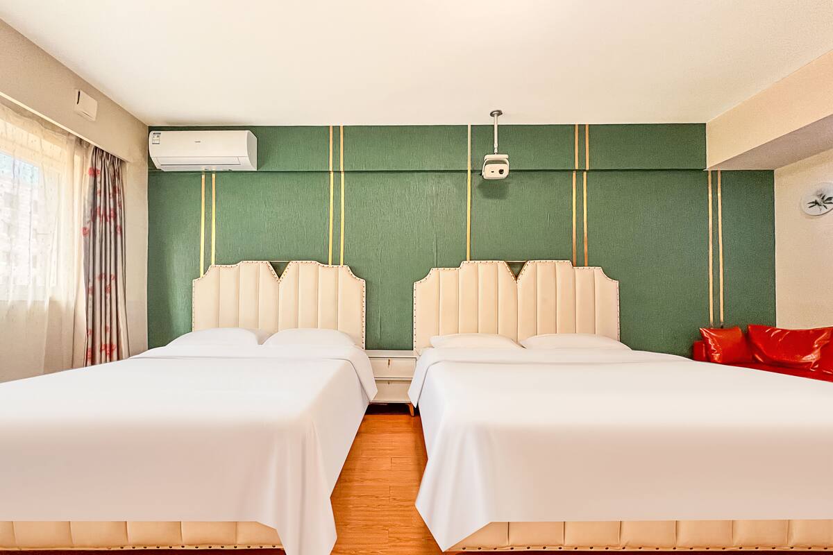 Habitación de hotel con dos camas, una pared de acento verde, suelo de madera, equipo de aire acondicionado y una silla roja.
