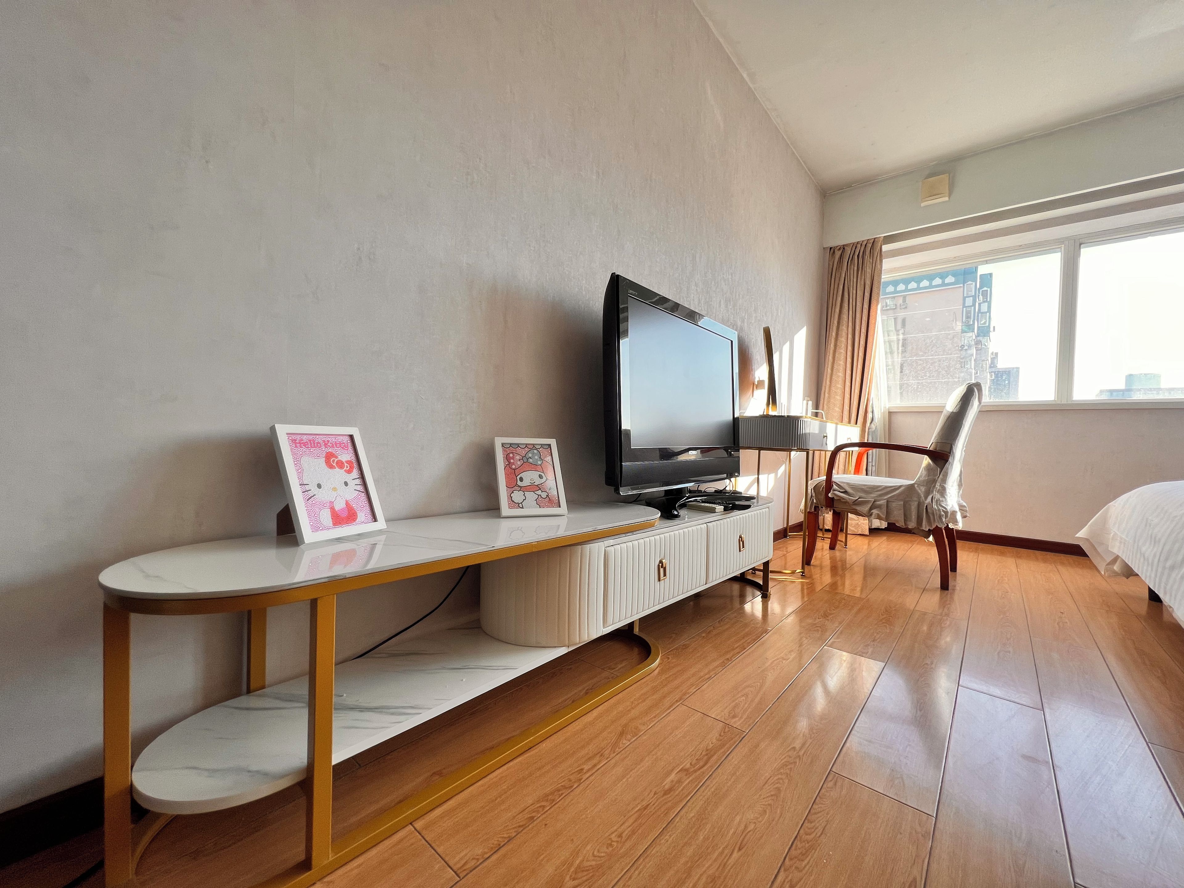 A hotel room with a TV on a white console, a bed, a chair, and a window with a city view.