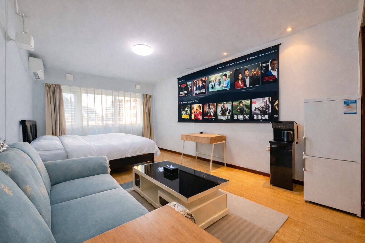 Habitación de hotel con cama, sofá, mesa de centro, escritorio pequeño, refrigerador y pared con carteles de películas.