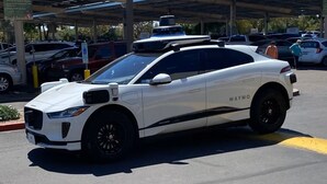 Ein weißes, autonomes Jaguar-Fahrzeug von Waymo steht auf einem Parkplatz. Im Hintergrund sind weitere Autos und eine überdachte Fläche zu sehen.