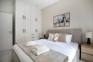 Ein ordentlich bezogenes Bett mit einem gemusterten Überwurf, ein großer Kleiderschrank mit mehreren Türen, ein Bild an der Wand und ein Nachttisch mit einer Lampe.