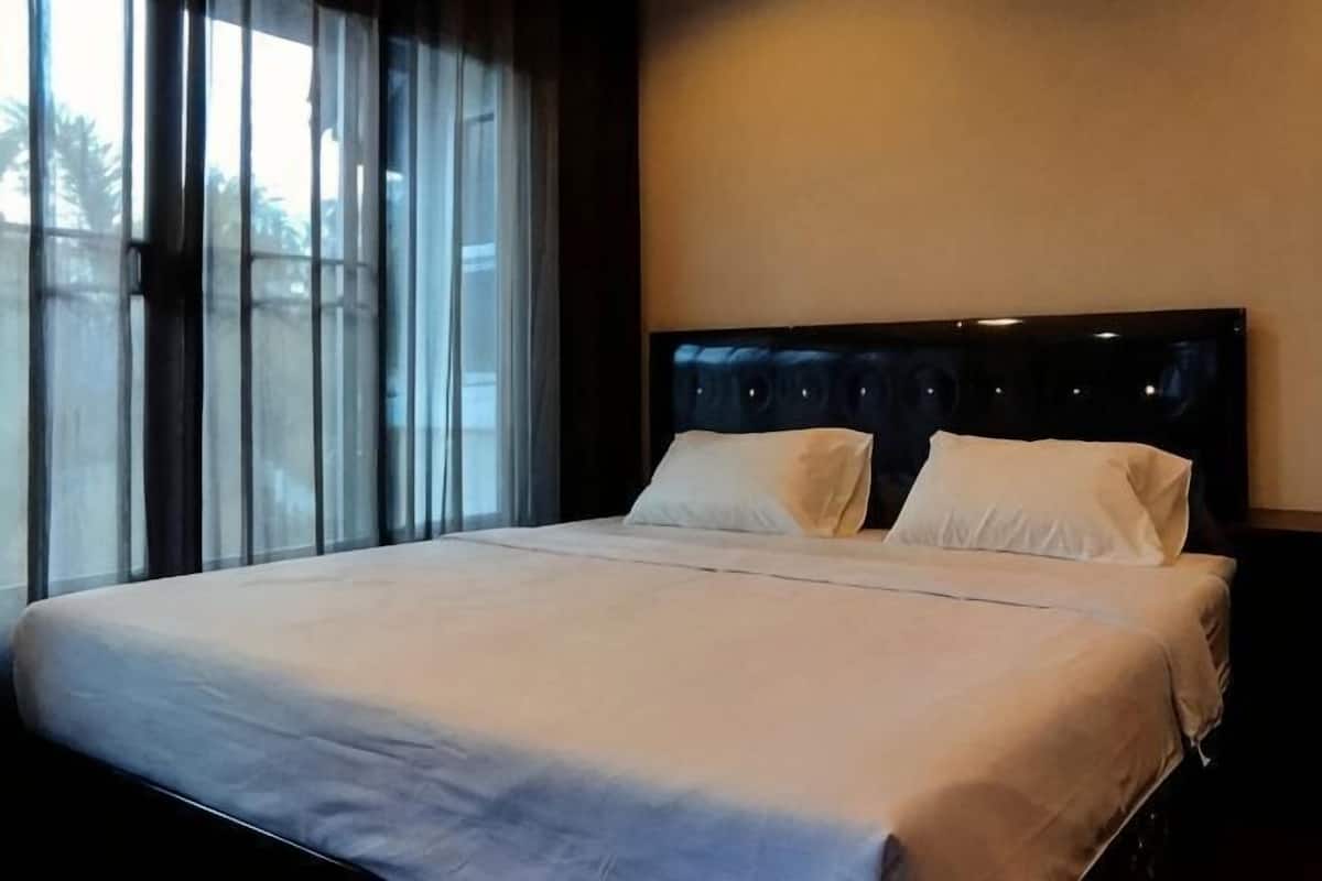 Ein ordentlich bezogenes Bett mit Kopfteil, ein Fenster mit Vorhängen und eine schlichte Wand.