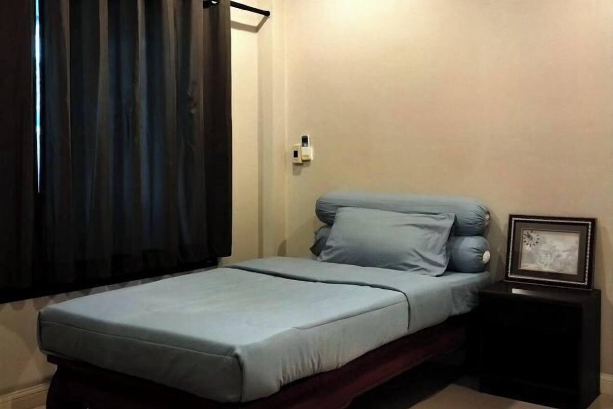 Ein Einzelbett mit blauer Matratze und Kissen, ein kleiner Nachttisch mit Bilderrahmen und eine Steckdose an der Wand.