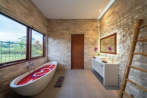Un baño con pared de piedra, puerta de madera, bañera con pétalos de flores, tocador blanco y un espejo.