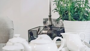 Een witte theepot, een witte kop, een witte pot met groene planten en een zwarte netcontainer op een wit oppervlak.