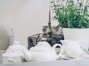 Ein weißer Teekessel, eine weiße Tasse, ein weißer Topf mit Grünpflanzen und ein schwarzer Netzbehälter auf einer weißen Oberfläche.