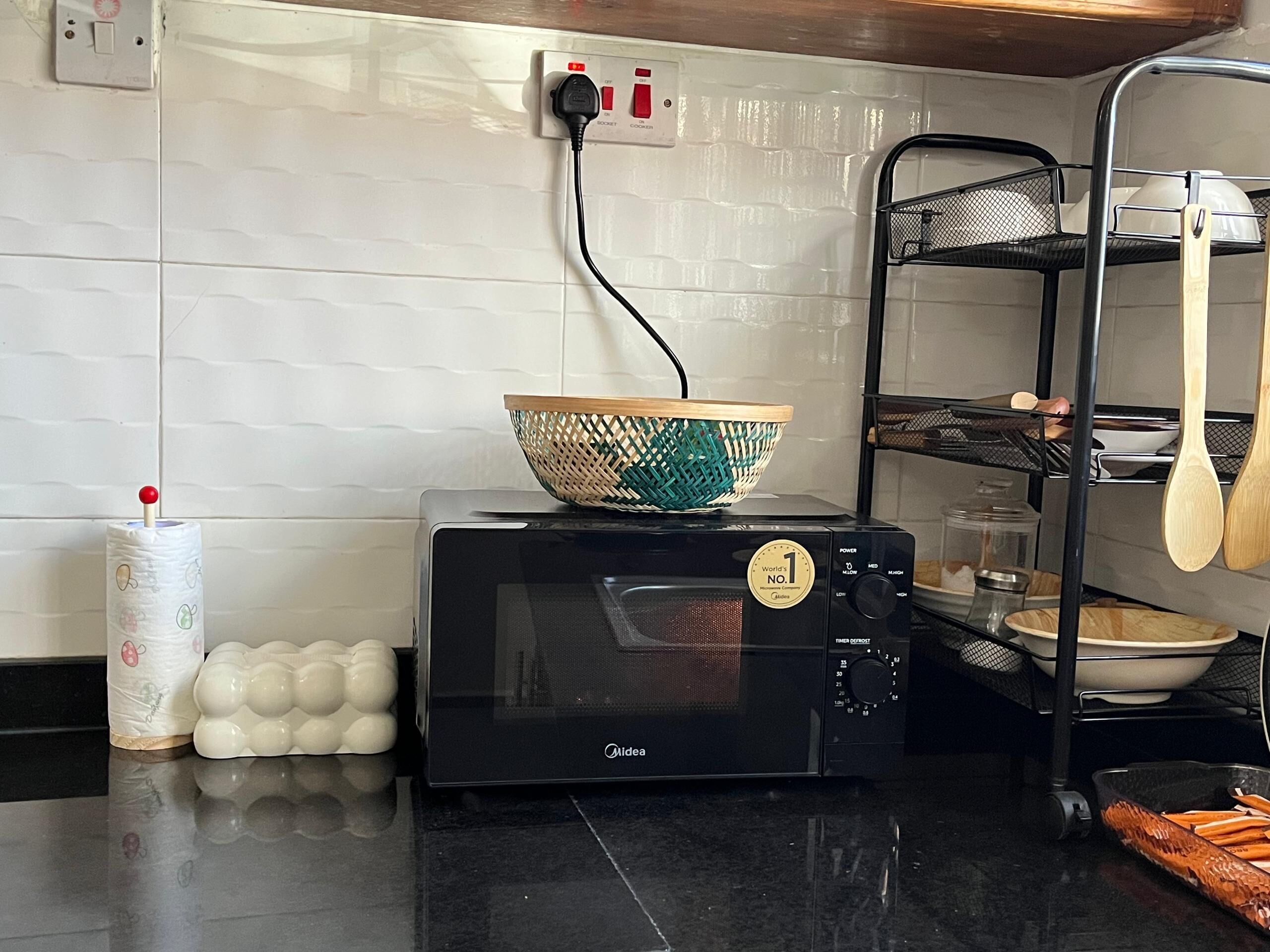 Uma cozinha com micro-ondas, tomada, cesta, rolo de papel toalha e um escorredor de louça.