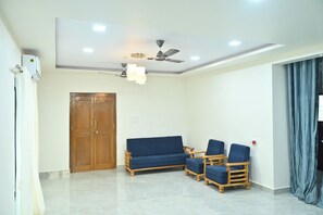 Una habitación con ventilador de techo, iluminación empotrada, una puerta de madera, un sofá azul y dos sillas de madera.