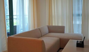 Una sala de estar moderna con un sofá seccional beige, una mesita con una caja de pañuelos y ventanas amplias con cortinas translúcidas.