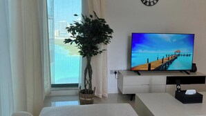 Un salón moderno con una televisión mostrando una escena de playa, una planta en maceta y vistas de un paisaje urbano a través de la ventana.