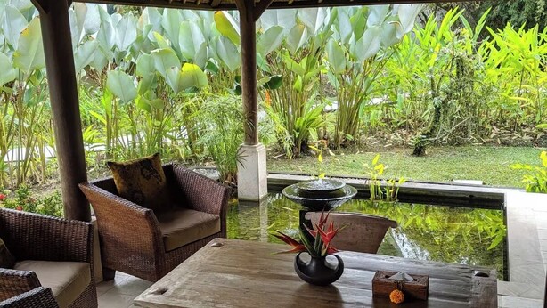 Un espacio de estar al aire libre con techo, amueblado con mobiliario de mimbre y una mesa central, con vistas a un jardín con diversas plantas y un elemento acuático.