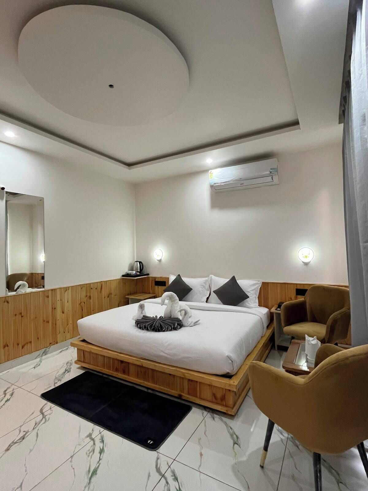 A hotel room with a bed, a wooden headboard, a small table, a chair, a mirror, and an air conditioning unit.