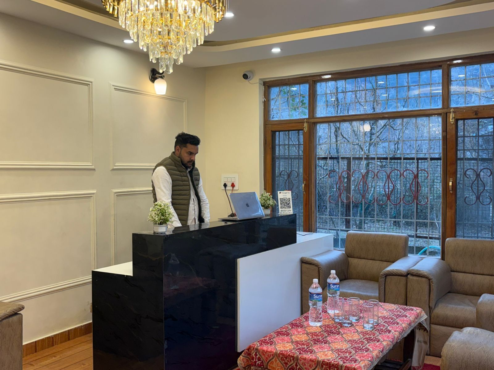 A hotel reception area with a marble counter, a chandelier, a large window with a view, and a table with water bottles.