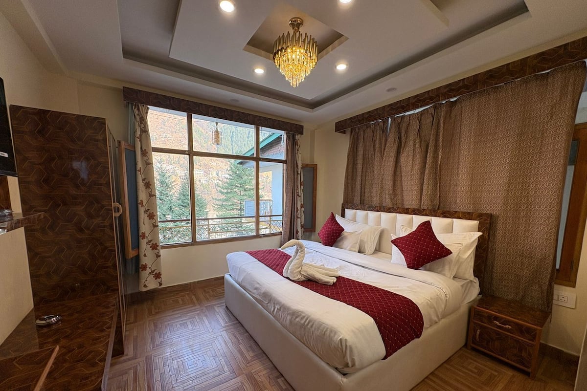 A hotel room with a large bed, a chandelier, a window with a view, and a wooden floor.