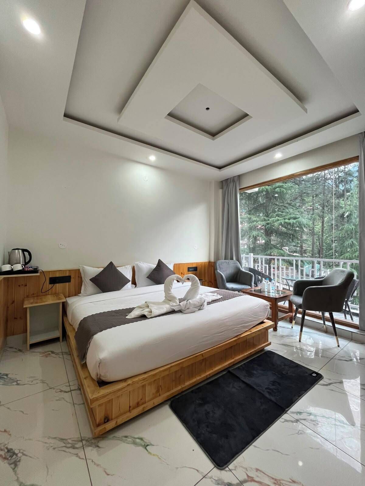 A modern hotel room with a large bed, a wooden headboard, a small desk with a kettle, a balcony with chairs, and a view of trees.