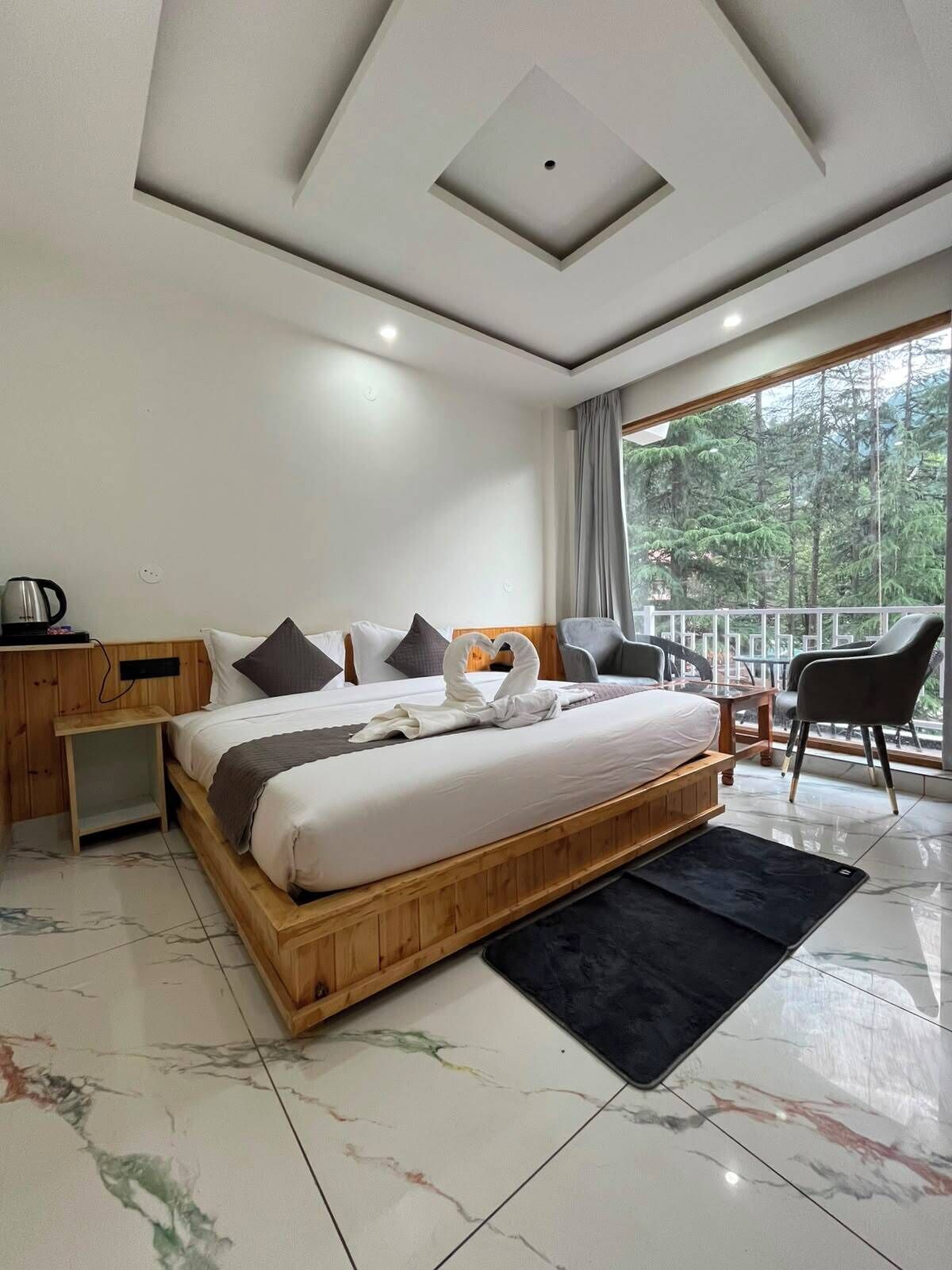 A modern bedroom with a wooden bed, a small bedside table, a chair, and a view of trees through large windows.