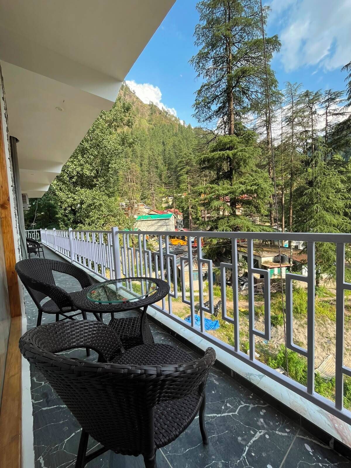 Un balcon doté de meubles en osier et d’une table en verre, offrant une vue sur une région montagneuse parsemée d’arbres et de bâtiments.