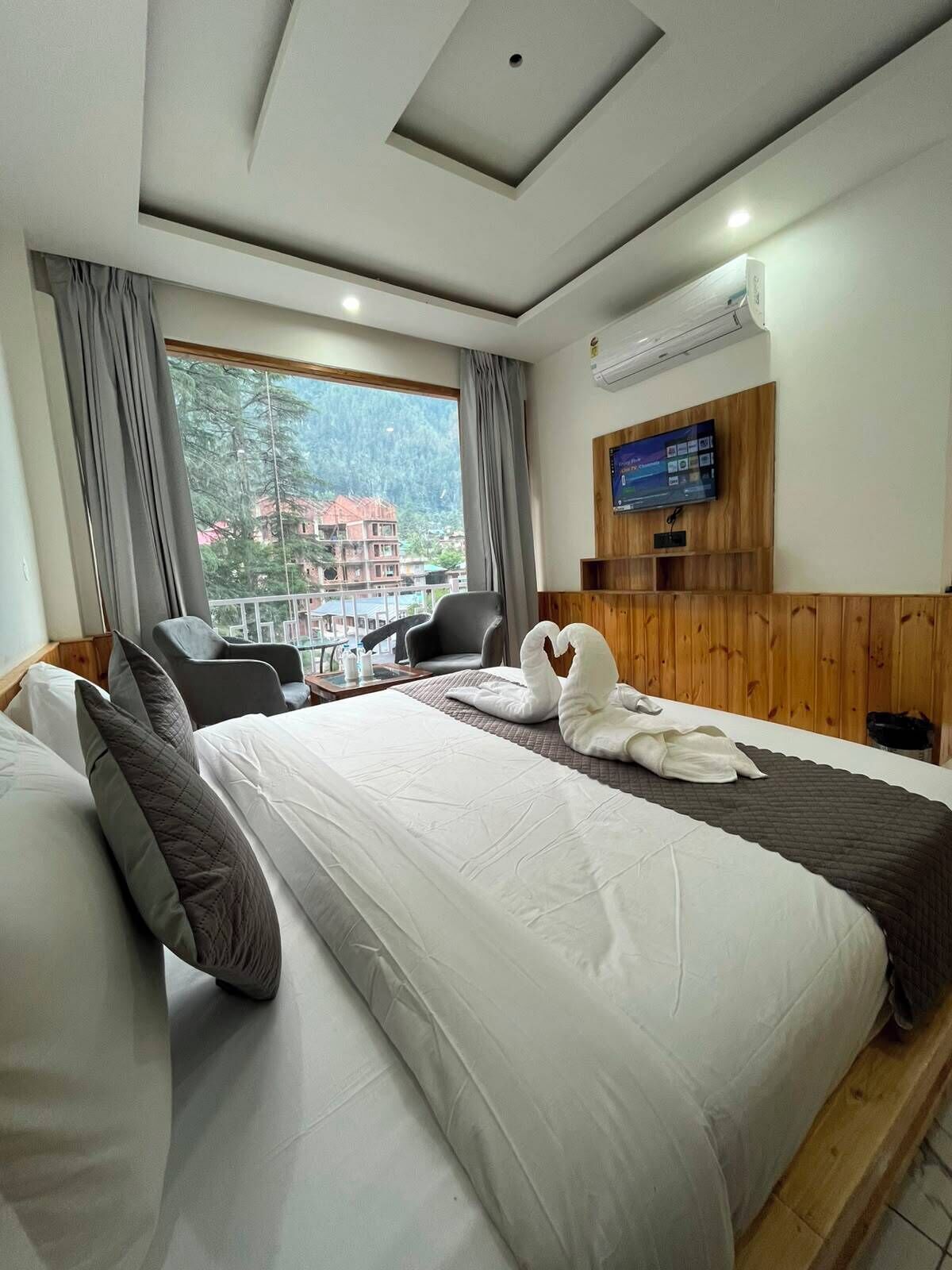 A hotel room with a large bed, two armchairs, a TV, and a view of a mountainous area.