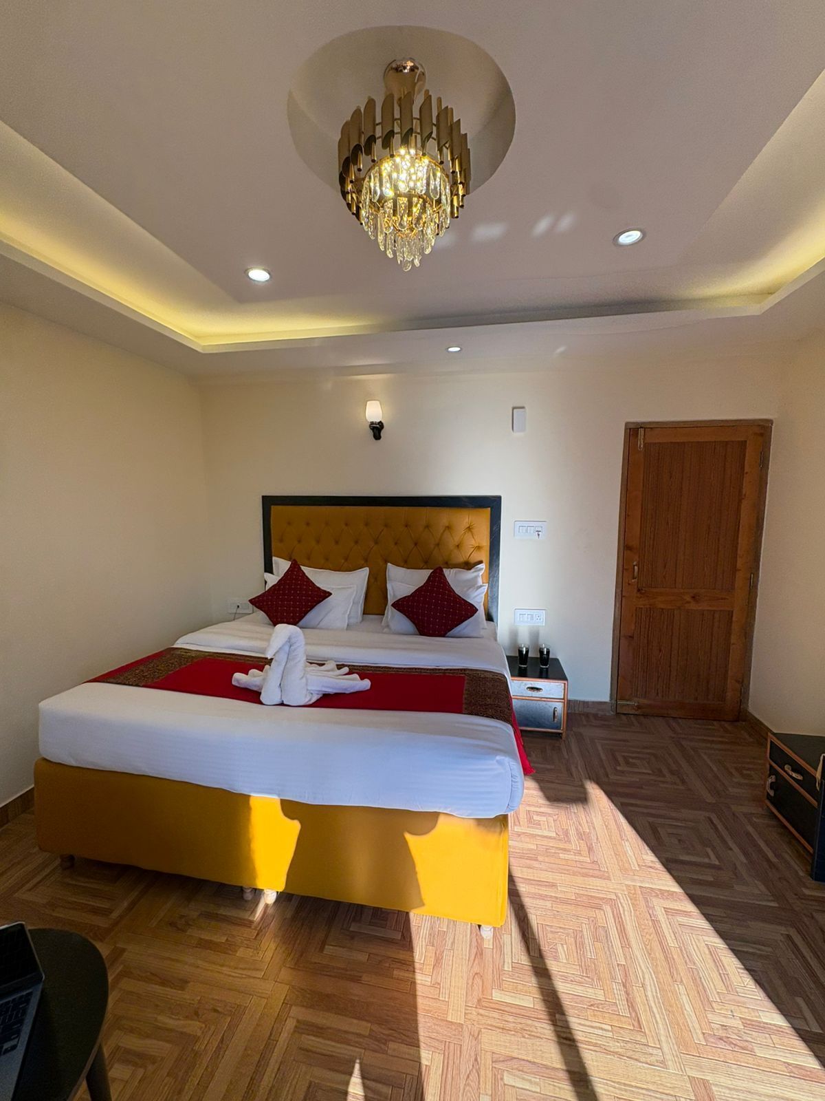 A hotel room with a large bed, a chandelier, a wooden door, and a nightstand with a lamp.