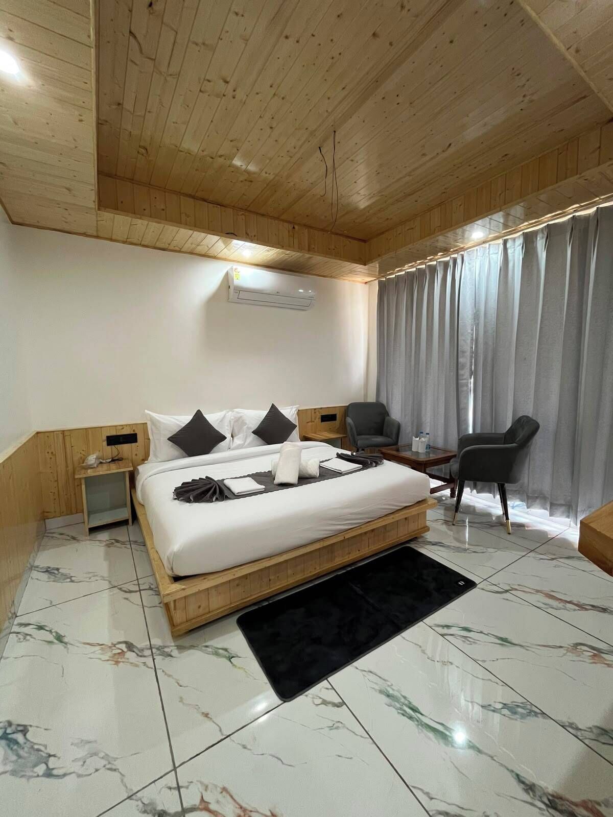 Habitación de hotel con techo de madera, cama con ropa blanca, un escritorio pequeño, una silla y una ventana con cortinas.