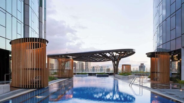 Una piscina en la azotea con estructuras cilíndricas de madera, un área de estar cubierta y edificios modernos al fondo.