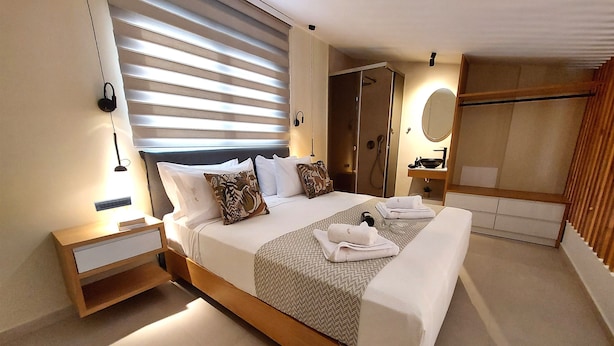 A modern hotel room with a large bed, bedside tables, a shower area, and a mirror. - Elea Suites Adelianos Kampos (Rethymno)