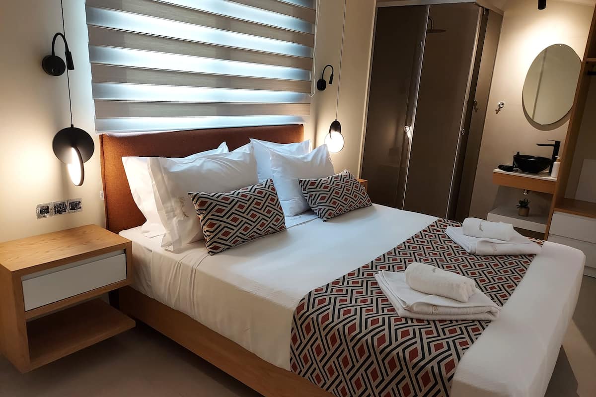 A neatly made bed with patterned bedding, a bedside table with a drawer, a nightstand, a wall-mounted lamp, and a mirror above the sink.