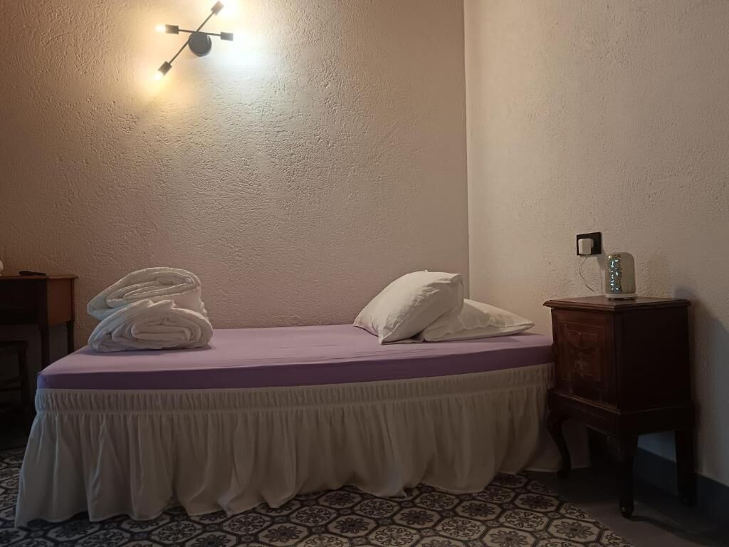 A room with a bed, a wall-mounted light fixture, a small wooden nightstand, and a patterned floor.