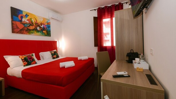 Una habitación de hotel con una cama roja, un escritorio con un control remoto, un televisor y un cuadro en la pared.