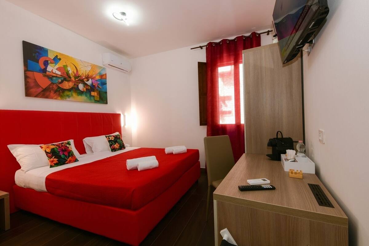 A hotel room with a red bed, a desk with a remote control, a TV, and a painting on the wall.