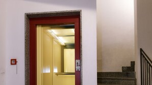 Un pasillo con una puerta de ascensor de marco rojo, un detector de incendios y una escalera de peldaños de granito.