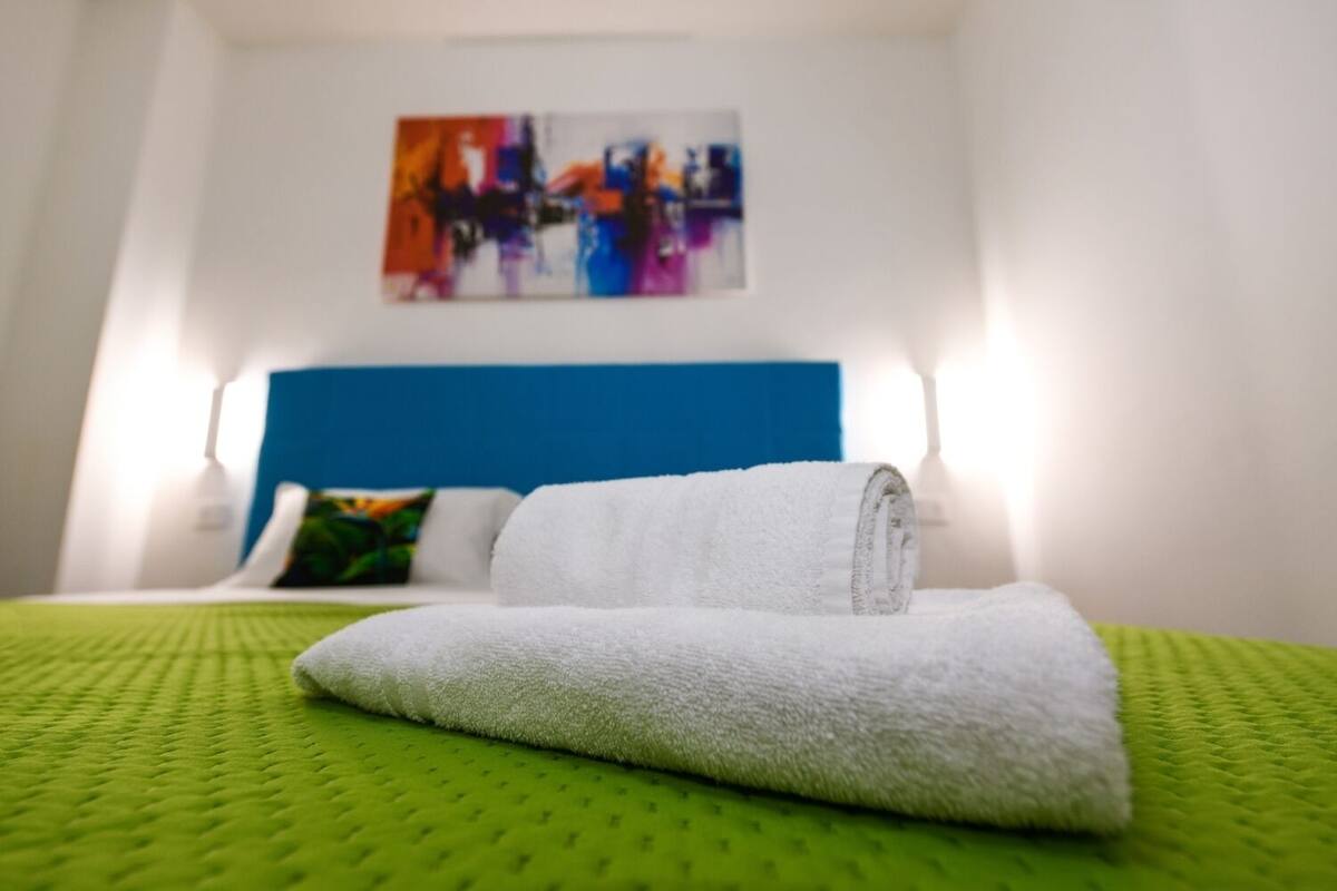 A hotel room with a bed, green bedspread, white towels, a blue headboard, and a colorful abstract painting on the wall.