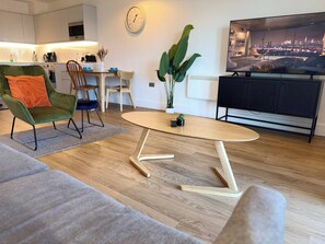 Ein modernes Wohnzimmer mit einem grünen Sessel, einem Holztisch, einem Essbereich mit Stühlen, einer Küche im Hintergrund und einer Fernsehansicht auf die Stadt.