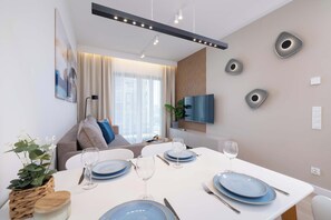 Un espace repas moderne avec une table blanche dressée pour quatre personnes, un canapé en arrière-plan et un téléviseur à écran plat fixé au mur.