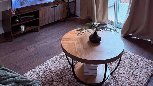 Una sala de estar con televisor, estantería, una mesa redonda de madera y un amplio ventanal con cortinas.