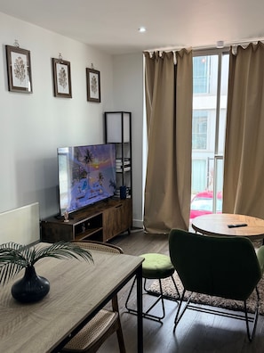 Ein modernes Wohnzimmer mit Flachbildfernseher, einem Esstisch mit Stühlen, einem Bücherregal und einem großen Fenster mit Vorhängen.